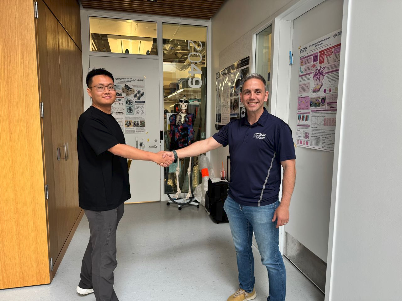 Photo in front of a lab with three academic posters behind two people smiling, looking at the camera, and shaking hands - left is Dr. Ang (Leo) Li and right is Dr. Mihai (Mishu) Duduta. 