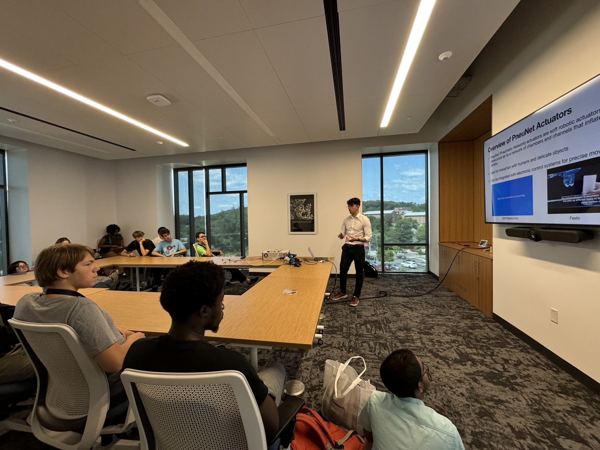 Lab alumni, Dom Flores, presents to students about Pneumatic Network Actuators (PneuNets) in a conference room. Students are seated at the conference tables with Dom standing at the front of the room presenting from a TV.