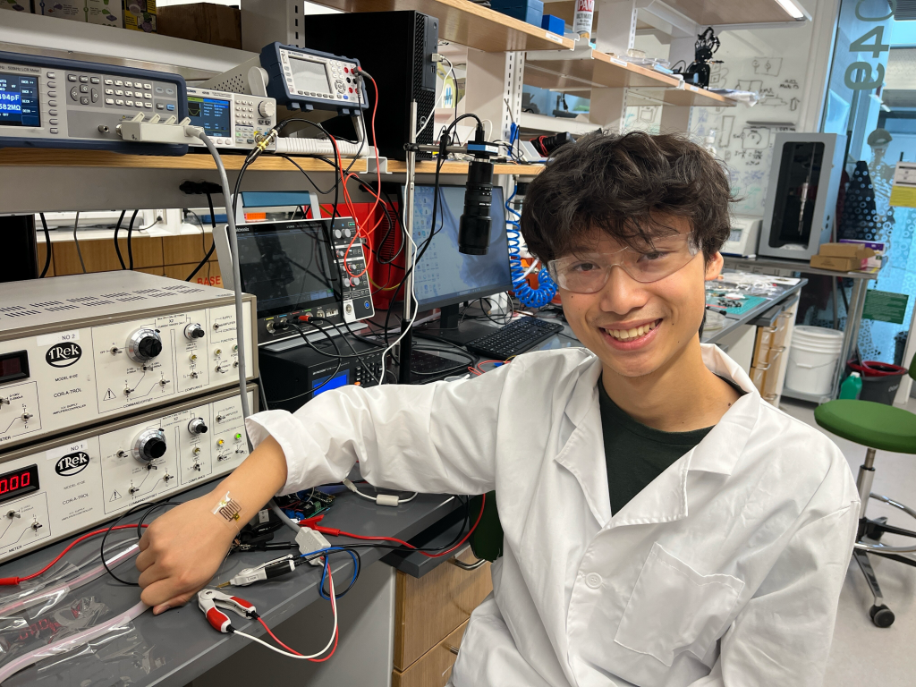 Hao Gu sitting at a lab bench in the Robot Incubator showing a soft strain sensor on his right hand.